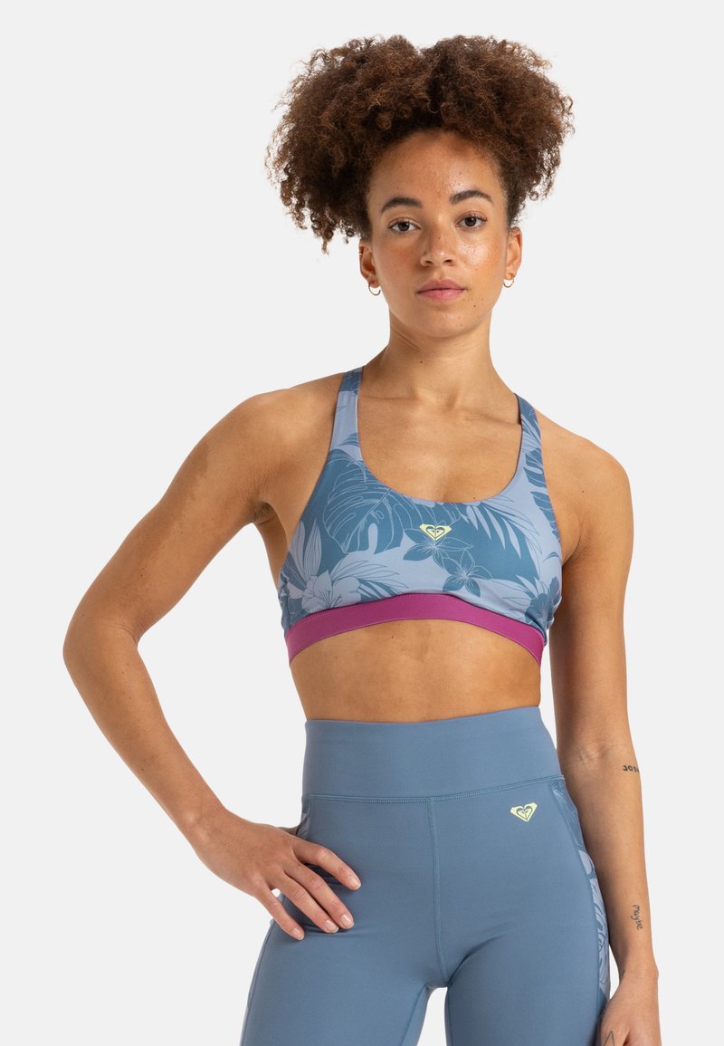 Sostenedor deportivo en azul con patrón de hojas tropicales, banda inferior púrpura y logo. Combinado con unos leggins ceñidos de cintura alta en azul a juego. Textura suave.