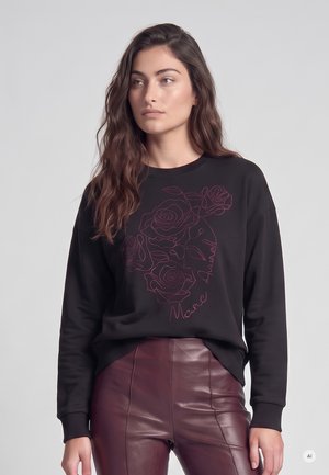 Junge Frau mit langen braunen Haaren, die einen schwarzen Pullover mit Rosenmotiven und bordeauxrote Lederhose trägt und zur Seite schaut.