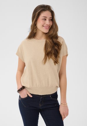 Ung kvinde med langt brunt hår iført beige kortærmet top, mørke jeans, guldringe, armbånd og ring, smilende og ser til siden.