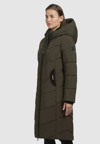 Dlouhý, tmavě zelený zateplený kabát s kapucí, který má diagonální prošívané vzory, přední zip a boční kapsy s černými akcenty.