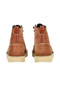 Stivali di pelle marrone con collare imbottito, suola in gomma e accenti arancioni a contrasto. Presenta il logo "SOREL" impresso sul tallone.
