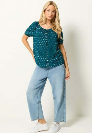 Blonde Frau steht mit Hand in der Hüfte, trägt eine türkisfarbene kurzärmelige Bluse mit weißen Punkten und hellblaue weite Jeans mit weißen Sneakers.