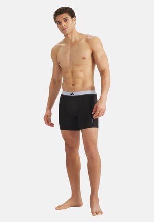 Oberkörperfreier Mann steht barfuß, trägt schwarze Boxershorts mit weißem Bund, auf dem ein schwarzes Adidas-Logo auf weißem Hintergrund zu sehen ist.