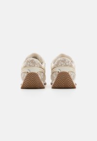 Zapatillas beige con parte superior de tejido estampado, detalles dorados y suela de goma texturizada con un patrón en zigzag en el talón.