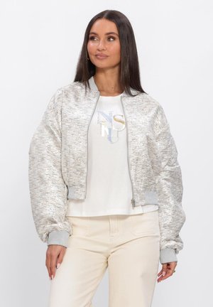 Vrouw met lang donker haar die een metalen zilverkleurige bomberjack draagt, een wit T-shirt met logo en lichtbeige broek, lichtjes opzij kijkend.