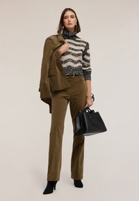 Hnědé kordurové kalhoty spojené se svetrem s černobílými pruhy a vysokým límcem. Drží černou tašku, na nohou má černé kotníkové boty.