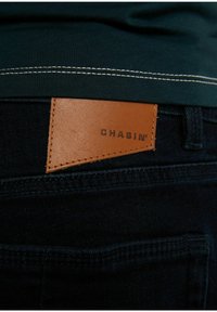 Vaqueros de denim negro con un parche de cuero marrón claro con "CHASIN'" grabado. Detalle de costura sutil por encima del bolsillo.