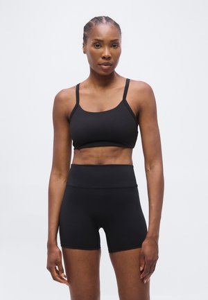 Fit Frau mit geflochtenem Haar trägt einen schwarzen Sport-BH und hoch taillierte schwarze Sportshorts, steht vor einem schlichten hellen Hintergrund.