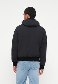 Veste à capuche noire avec texture rembourrée, poignets et ourlet côtelés, conçue dans une coupe décontractée, présentée de dos sur fond blanc.