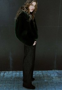 Modelka w ciemnozielonym, puszystym, oversize'owym swetrze i prostych czarnych dżinsach, stojąca na tle ciemnego tła.