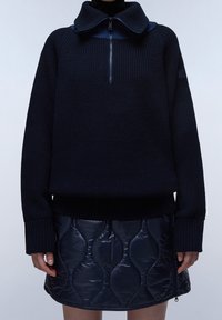 Granatowy sweter z dzianiny z kołnierzykiem na zamek, w połączeniu z pikowaną granatową spódnicą z owalnym wzorem i zasuwaną na boku detalem.