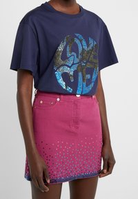 Camiseta de algodón color azul marino con un gran gráfico de lentejuelas que dice "AMAME". Combinada con una falda de mezclilla rosa con acentos de puntos azules y negros en el dobladillo.