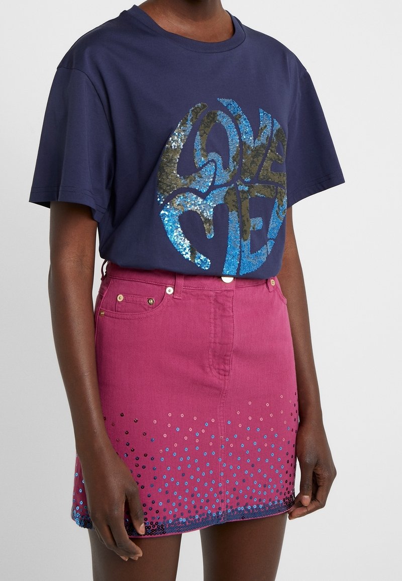 Camiseta de algodón color azul marino con un gran gráfico de lentejuelas que dice "AMAME". Combinada con una falda de mezclilla rosa con acentos de puntos azules y negros en el dobladillo.