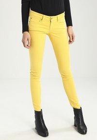 Persona con pantalones amarillos ajustados, camiseta de manga larga negra y botas negras hasta el tobillo, de pie sobre un fondo blanco.