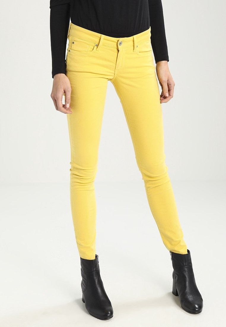 Persona con pantalones amarillos ajustados, camiseta de manga larga negra y botas negras hasta el tobillo, de pie sobre un fondo blanco.