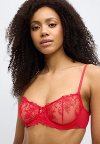 Žena kovrčave kose koja nosi crveni čipkasti grudnjak s cvjetnim vezom i malom mašnom sprijeda, na jednobojnoj svijetloj pozadini.