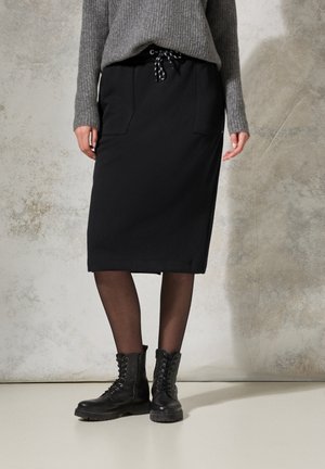 Zwarte rok tot de knie met twee voorzakken en een trekkoord in de taille. Gecombineerd met zwarte veterschoenen en een grijze gebreide trui.