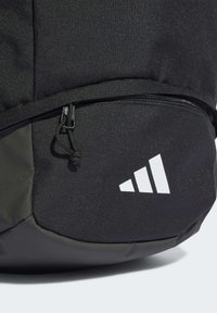 Schwarzer Rucksack mit strukturierter Oberfläche, einem Reißverschlussfach und einem weißen Logo mit drei Streifen. Der Boden hat eine glatte, dunkelgrüne Oberfläche.