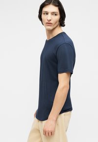 Giovane uomo con capelli scuri fino alle spalle, che indossa una maglietta a maniche corte blu navy e pantaloni beige, che guarda di lato.