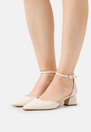 Zapatos con tacón bloque en beige claro, con punta afilada y correa en el tobillo. Fabricados en un material suave, presentan un diseño de escote bajo y acentos mínimos.