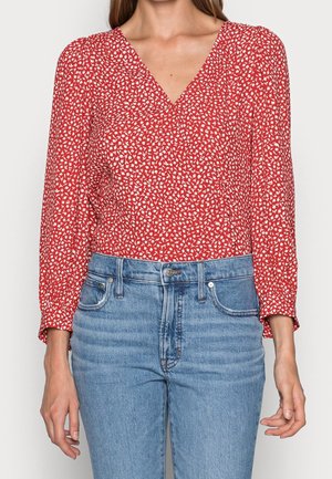 Kvinde iført en rød langærmet V-halsbluse med hvidt blomsterprint og lyseblå jeans med høj talje, stående med afslappede arme.