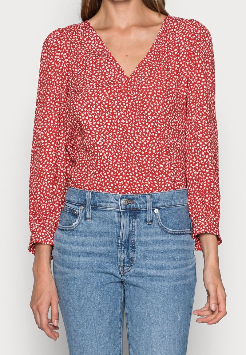 Femme portant un chemisier rouge à manches longues col en V avec imprimé floral blanc et un jean bleu clair taille haute, debout les bras détendus.