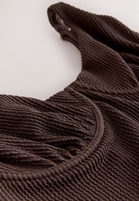 Primo piano di un top in tessuto marrone scuro con dettagli arricciati e design senza maniche su uno sfondo chiaro.
