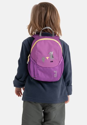 Bambino con capelli castano chiaro fino alle spalle che indossa una giacca blu navy e pantaloni grigi, con uno zaino viola decorato con cartoni animati di animali.