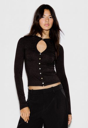 Femme aux longs cheveux foncés portant un haut noir ajusté à manches longues avec une découpe devant et des boutons blancs, associé à un pantalon noir.