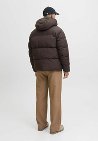 Man in een donkerbruine pufferjack met capuchon, beige broek, beige schoenen en een zwarte pet, van achteren gezien op een witte achtergrond.