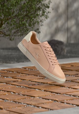 Világos rózsaszín Calvin Klein sportcipő fehér talppal és sarokkal, amely egy kültéri kő- és fa textúrájú talaj fölött lebeg.