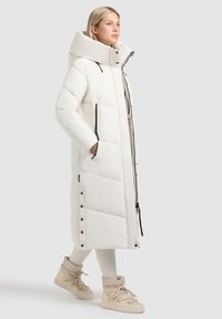 Ilgas baltas pūkinis paltas su gobtuvu, pasižymintis siuvinėjimo dizainu, dviem užtrauktukais uždaromomis kišenėmis ir mygtukų akcentais šone.