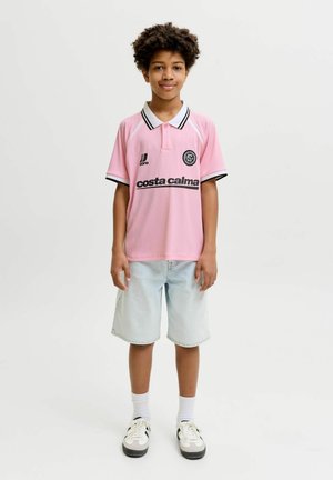 Băiatul stă cu fața spre înainte purtând un tricou polo roz cu text negru, pantaloni scurți de denim deschis, șosete albe și adidași alb-negru pe un fundal alb.