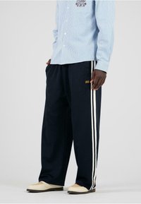 Marineblaue weit geschnittene Hose mit weißen Seitenstreifen, aus weichem Stoff gefertigt. Kombiniert mit einem hellblauen gestreiften Hemd mit Knopfleiste.