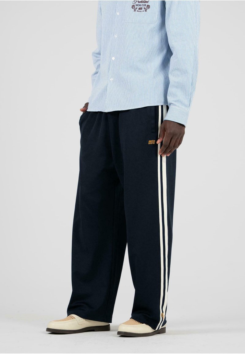 Marineblaue weit geschnittene Hose mit weißen Seitenstreifen, aus weichem Stoff gefertigt. Kombiniert mit einem hellblauen gestreiften Hemd mit Knopfleiste.