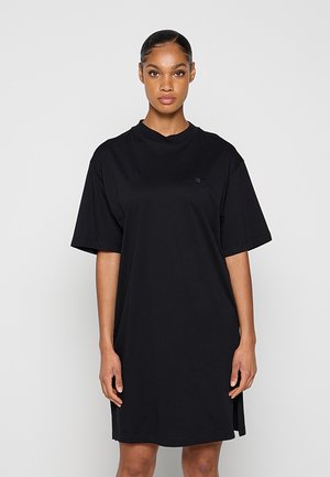 Vrouw draagt een effen zwarte oversized T-shirtjurk, staand tegen een lichtgrijze achtergrond, met een neutrale uitdrukking recht vooruit kijkend.