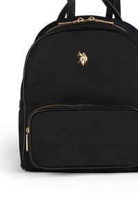 Svart nylonryggsäck med guldfärgad dragkedja och metallbeslag, rundad design, framficka och en liten logotyp på den övre delen.