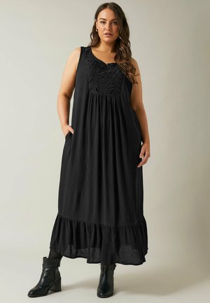 Sleeveless black maxi dress with embroidered bodice, gathered waist, and tiered hem. Smooth texture, side pockets, paired with black ankle boots.