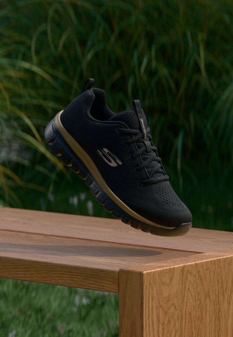 Baskets Skechers noires avec des accents dorés et une semelle robuste flottant au-dessus d’un banc en bois, herbe verte et plantes en arrière-plan.