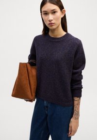 Dunkellila gestrickter Pullover mit geripptem Kragen, kombiniert mit hellblauen Jeans. Beigefarbenes Wildlederhandtasche in der rechten Hand gehalten.