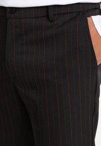 Persoon die zwarte broek draagt met dunne rode verticale strepen en een wit overhemd, hand casual in de zak.