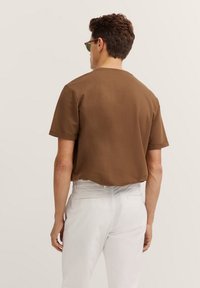 Braunes Baumwoll-T-Shirt mit kurzen Ärmeln, lockerer Passform und Rundhalsausschnitt, kombiniert mit weißen Hosen. Sauberes Design, glatte Textur.