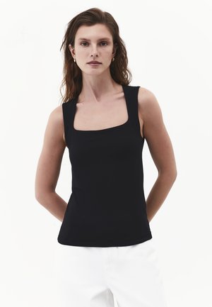Frau mit welligem braunem Haar, die goldene Creolen, ein schwarzes ärmelloses Top mit quadratischem Ausschnitt und weiße Hosen trägt, steht vor einem schlichten Hintergrund.