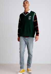 Gilet verde a coste con una lettera "C" bianca, indossato sopra una maglietta bordeaux a righe, abbinato a jeans azzurri e sneakers gialle.