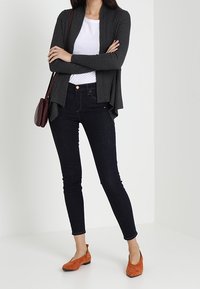 Grå ribbad kofta över en vit T-shirt, kombinerad med mörka tajta jeans och orange mockaskor. Inkluderar en liten vinröd handväska.