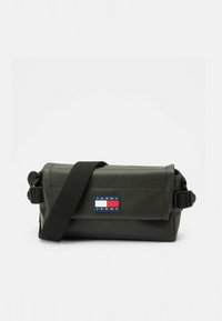 Pravokutna tamnozelena torba od sjajnog materijala. Ima podesivi remen i naljepnicu s logotipom Tommy Jeans u crvenoj, bijeloj i plavoj boji.