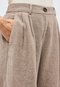 Μπεζ λινό παντελόνι με πιέτες, ζώνη με κουμπιά, θηλιές για ζώνη και ένα χέρι στην αριστερή τσέπη.