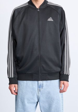 Personne portant une veste de survêtement noire Adidas avec des bandes blanches sur les manches et une fermeture éclair à l'avant, associée à un jean bleu clair.