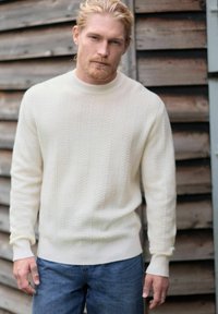 Pull en tricot de couleur crème avec un motif en chevron texturé, col et poignets côtelés, associé à un jean en denim bleu.