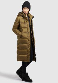 Manteau matelassé long vert olive avec capuche, doté d'une fermeture éclair et de poches, associé à des leggings noirs et des bottes de cheville noires en peluche.